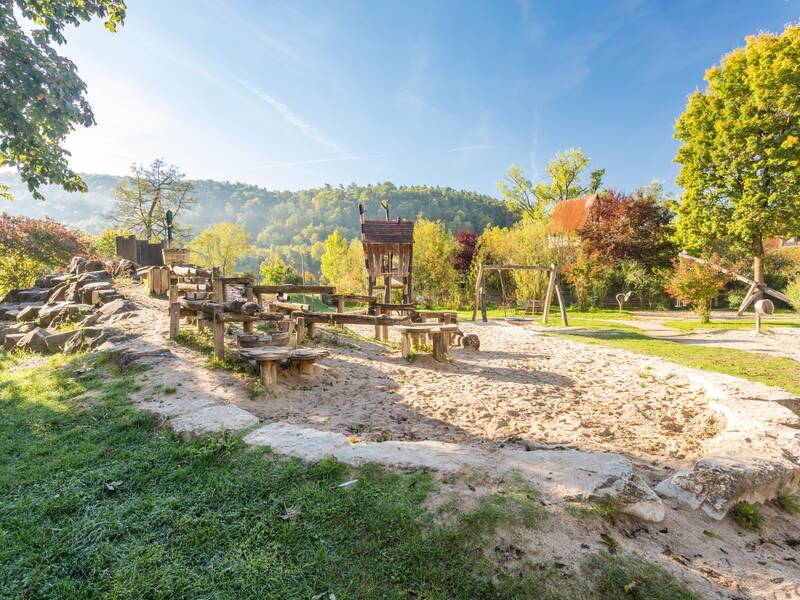 Ein idyllischer Spielplatz mit Sandfläche und Holzkonstruktionen ist von Bäumen und Hügeln umgeben, unter strahlend blauem Himmel.