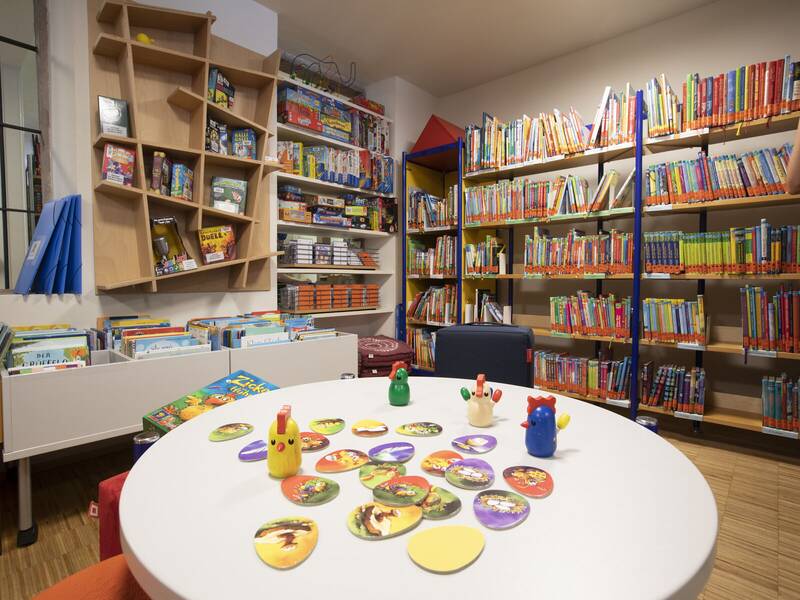 Ein Raum mit einem großen Tisch, auf dem Spielsteine liegen, umgeben von Regalen voller Bücher und Spiele.