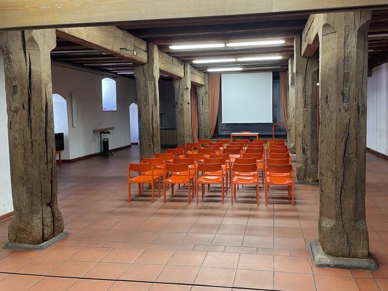 Ein Konferenzraum mit Holzbalken, einer großen Leinwand und mehreren Reihen roter Stühle.