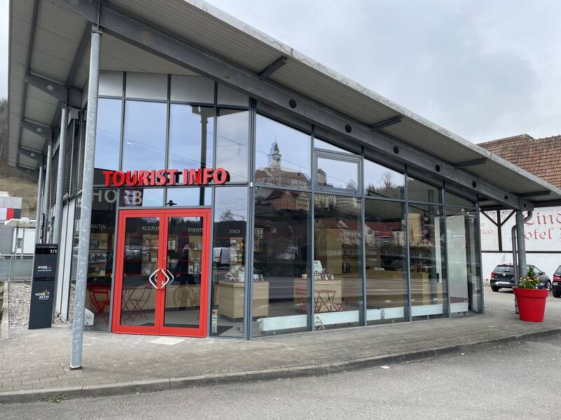 Das Bild zeigt ein modernes Gebäude mit Glasfassade und roter Tür, das als Tourist-Info dient.