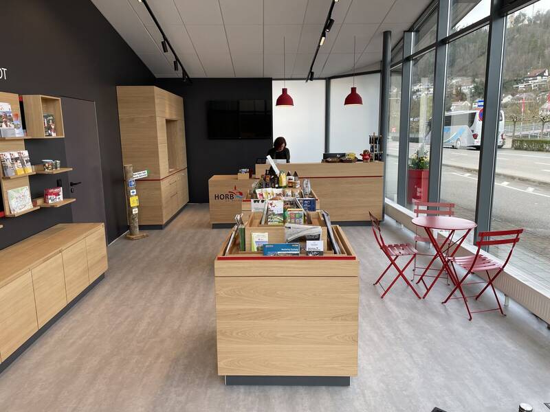 Das Bild zeigt einen Raum mit Holzmöbeln, auf denen Prospekte ausgestellt werden. Im Hintergrund ist eine Rezeption zu sehen, an der eine Person arbeitet.