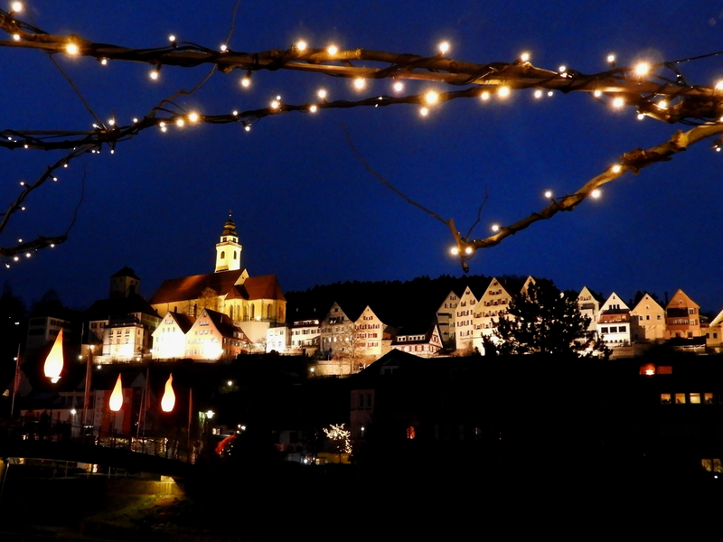Eine nächtliche Szenerie zeigt eine beleuchtete Stadt mit historischen Gebäuden und einer Kirche im Hintergrund, eingerahmt von Lichterketten im Vordergrund.