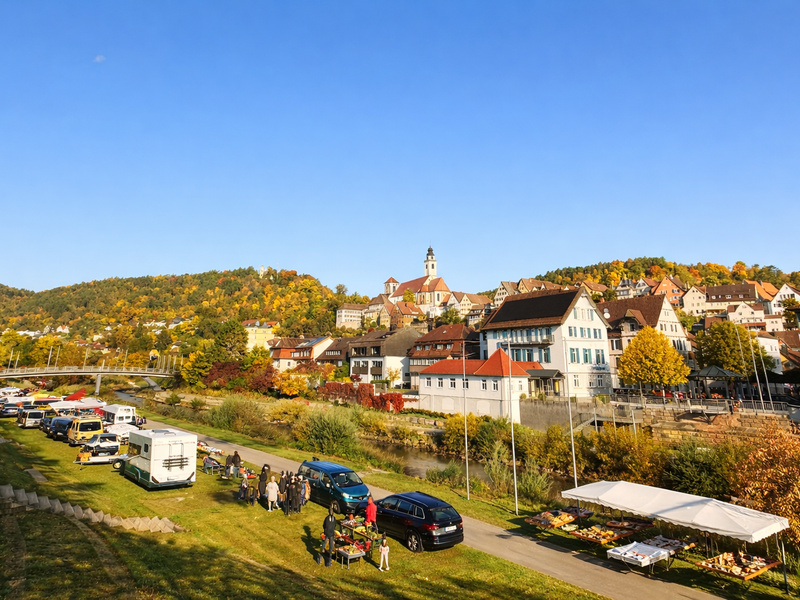 Der Flohmarkt von Horb am Neckar am Ufer des Neckars, mit Marktständen, Besuchern und der historischen Stadtsilhouette mit Kirche im Hintergrund.