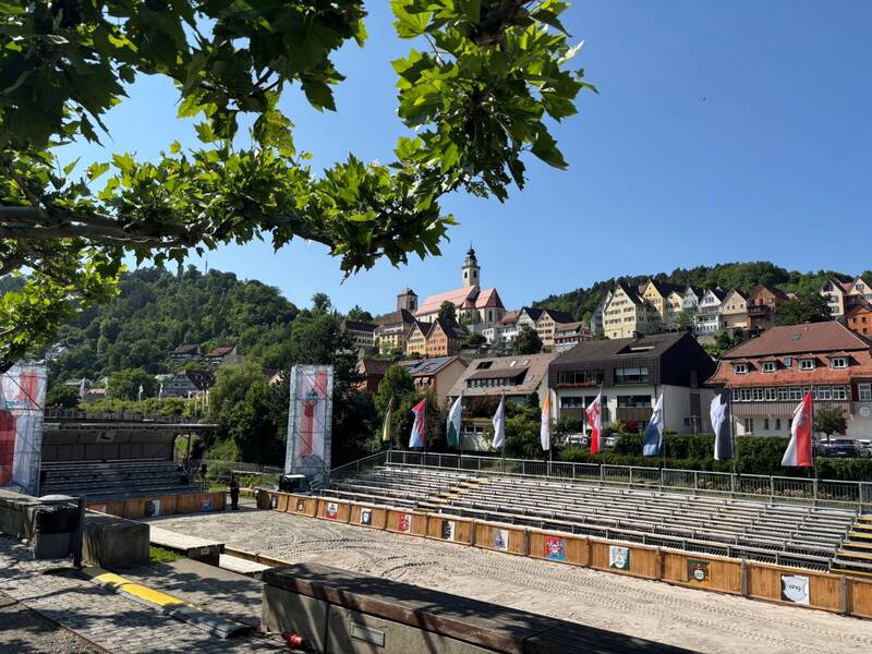 Die Aufnahme zeigt eine Freilichtbühne mit leeren Sitzreihen, flankiert von Bäumen, vor einer malerischen Kulisse aus bunten Häusern und einer Kirche auf einem Hügel, unter klarem, blauem Himmel.
