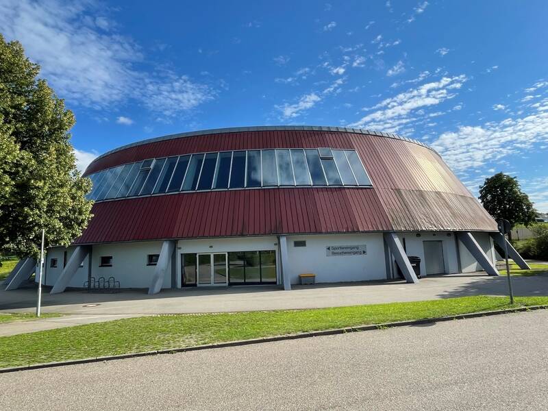 Das Bild zeigt ein großes, rundes Gebäude mit einer rotbraunen Metallfassade, Fenstern im oberen Bereich und einem angrenzenden Gehweg, umgeben von Bäumen, unter einem blauen Himmel.