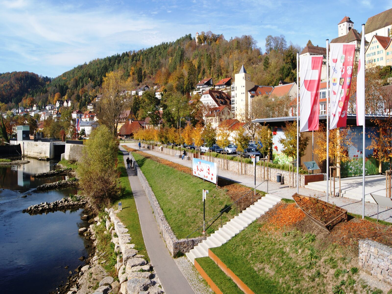 Eine malerische Neckarlandschaft mit einem Uferweg, herbstlich gefärbten Bäumen, mehreren Flaggen und im Hintergrund der Horber Flößerweg an einem bewaldeten Hang.