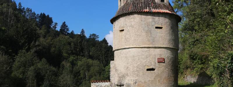Ein runder, historischer Turm mit einem steilen, spitzen Dach steht vor einem Hintergrund aus dicht bewaldeten Hügeln und blauem Himmel.