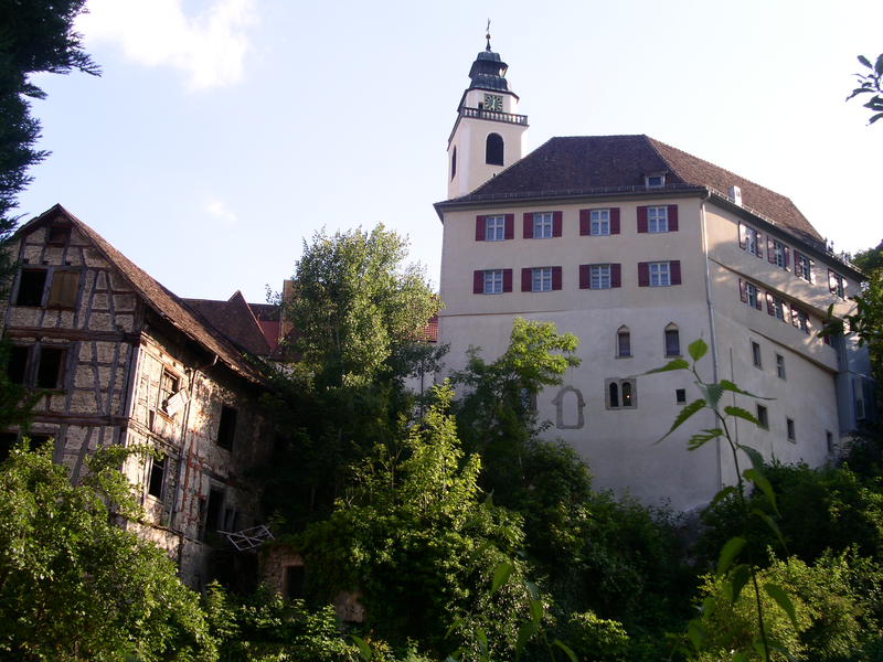 Ein großes, mehrstöckiges Gebäude mit rotem Dach und roten Fensterläden steht neben einem älteren Fachwerkhaus, umgeben von üppiger Vegetation.