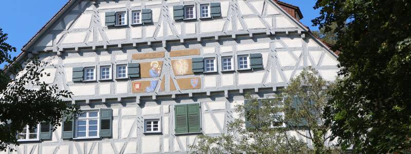 Ein traditionelles Fachwerkgebäude mit mehreren Fenstern und grünen Fensterläden, umgeben von sommerlichem Blattwerk.