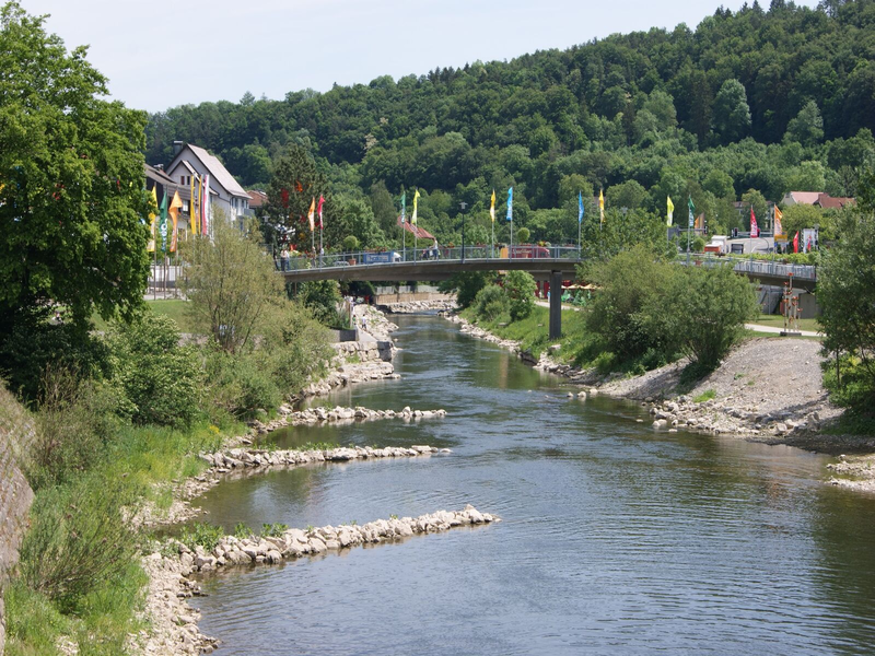 Ein kleiner Fluss fließt unter einer Brücke hindurch, umgeben von grünen Bäumen und Fahnen auf beiden Seiten.