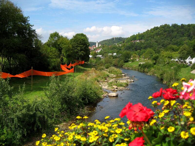 Eine malerische Stadtansicht mit traditionellen Häusern und einer Kirche spiegelt sich in einem ruhigen Fluss wider, umgeben von grüner Landschaft.