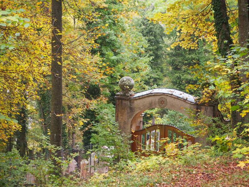 Ein Torbogen mit einem Davidstern führt zu einem verwunschenen Friedhof, umgeben von herbstlichem Wald.