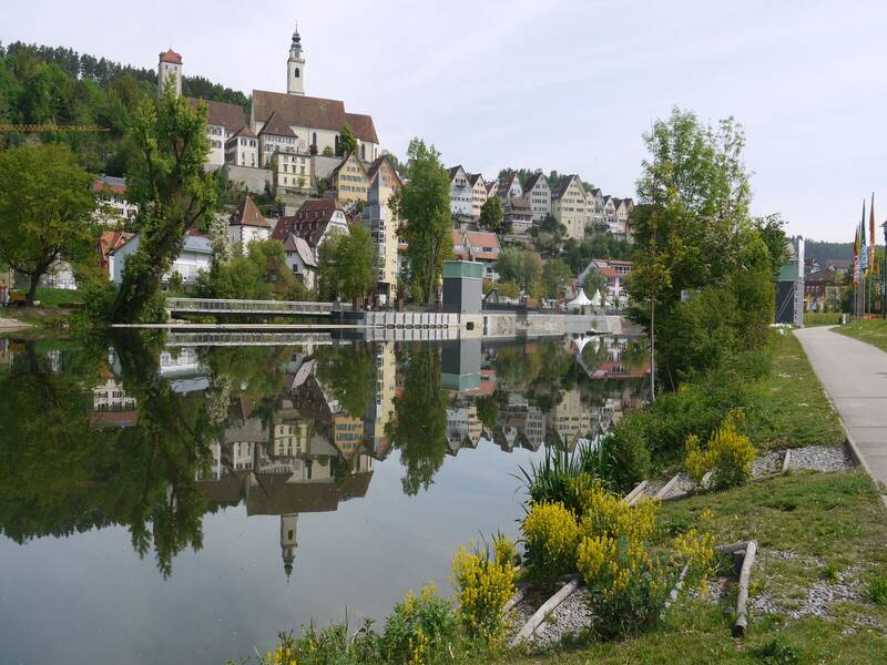 Eine malerische Stadtansicht mit einer Kirche und bunten Häusern, die sich in einem ruhigen Fluss spiegeln, umgeben von grünen Bäumen und einem Gehweg am Ufer.