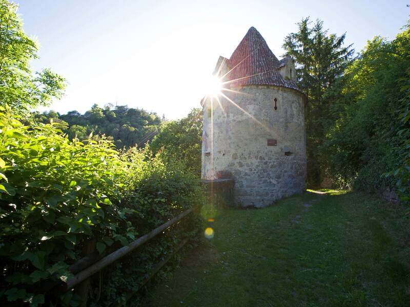 Ein runder, steinerner Turm mit einem roten Dach steht inmitten grüner Vegetation, während die Sonne dahinter aufgeht.