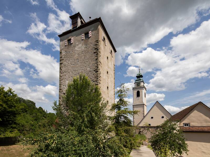 Das Bild zeigt einen historischen Steinturm und einen Kirchturm im Hintergrund, umgeben von Bäumen, unter einem teils bewölkten Himmel.