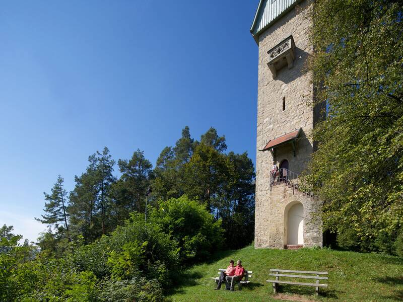 Zwei Personen sitzen auf einer Bank vor einem steinernen Turm, der von Bäumen umgeben ist und in den blauen Himmel ragt.