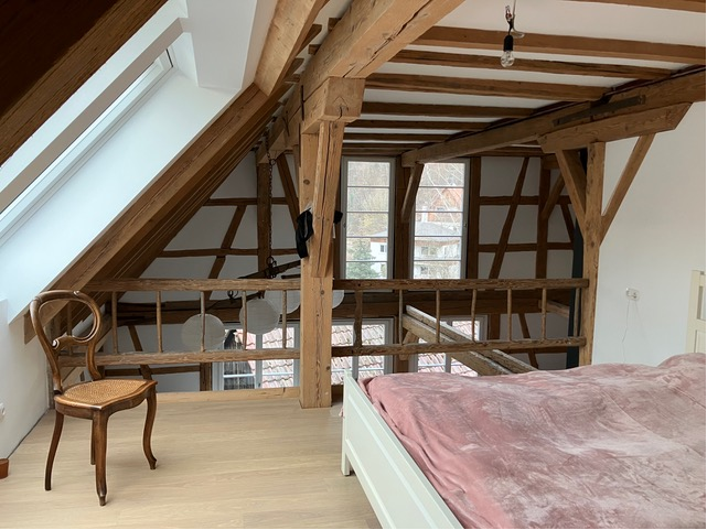 Das Bild zeigt ein gemütliches Dachzimmer mit sichtbaren Holzbalken, einem Stuhl, einem Bett mit rosafarbener Decke und einem großen Fenster im Hintergrund.