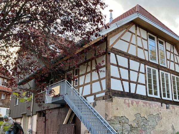 Ein traditionelles Fachwerkhaus mit großflächigen Fenstern, einer Außentreppe und herbstlichem Laub im Vordergrund.