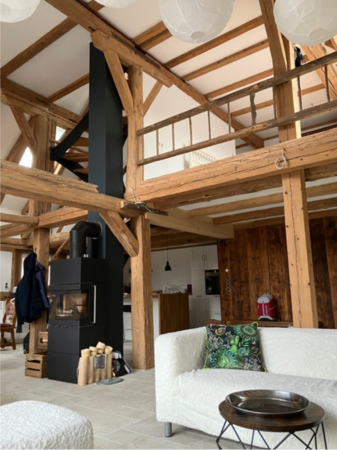 Ein offener Wohnraum mit sichtbaren Holzbalken, einem schwarzen Kaminofen und einem weißen Sofa mit dekorativem Kissen.