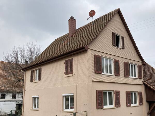 Das Bild zeigt ein zweistöckiges Haus mit einem Satteldach, mehreren Fenstern mit Holzläden und einer Satellitenschüssel auf dem Dach.