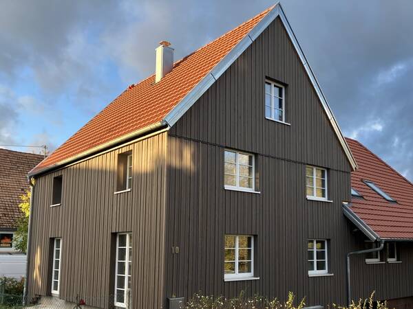 Ein zweistöckiges Haus mit dunkelbrauner Holzfassade und rotem Satteldach steht unter einem bewölkten Himmel.
