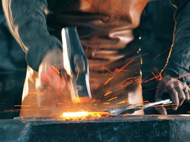 Ein Schmied hämmert mit einem großen Hammer auf ein glühendes Metallstück auf einem Amboss, wobei Funken sprühen.
