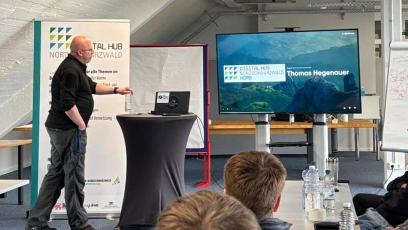 Das Bild zeigt einen Mann der eine Präsentation hält. Der Laptop steht auf einem Stehtisch und zeigt die Präsentation auf einem Fernseher. Hinter dem Mann steht ein Roll-Up Banner. Im Vordergrund sind die Hinterköpfe von Zuschauenden und ein Tisch mit Wasserfalschen zu sehen. 