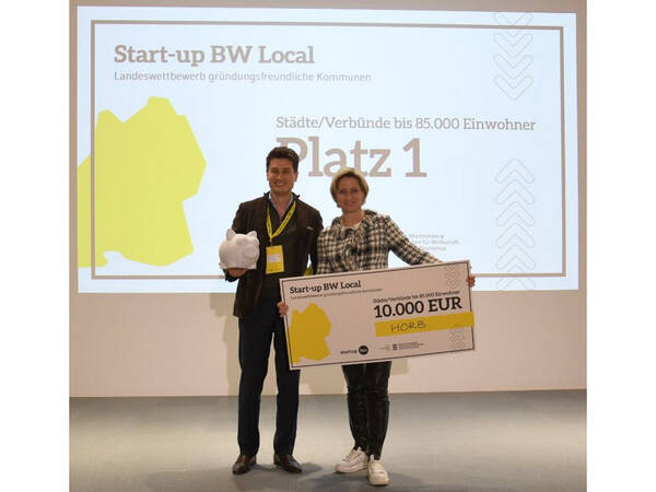 Zwei Personen stehen auf einer Bühne und halten einen großen Scheck über 10.000 Euro im Rahmen des Wettbewerbs "Start-up BW Local", während eine Projektionsfolie im Hintergrund den ersten Platz anzeigt.