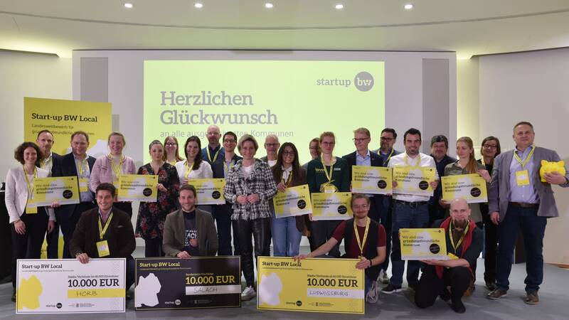 Eine Gruppe von Menschen hält auf einer Bühne verschiedene große Schecks und Schilder, während sie für ein Gruppenfoto vor einem gelb-weißen Hintergrund posiert, auf dem "Herzlichen Glückwunsch" steht.