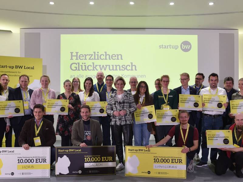 Eine Gruppe von Menschen hält auf einer Bühne verschiedene große Schecks und Schilder, während sie für ein Gruppenfoto vor einem gelb-weißen Hintergrund posiert, auf dem "Herzlichen Glückwunsch" steht.