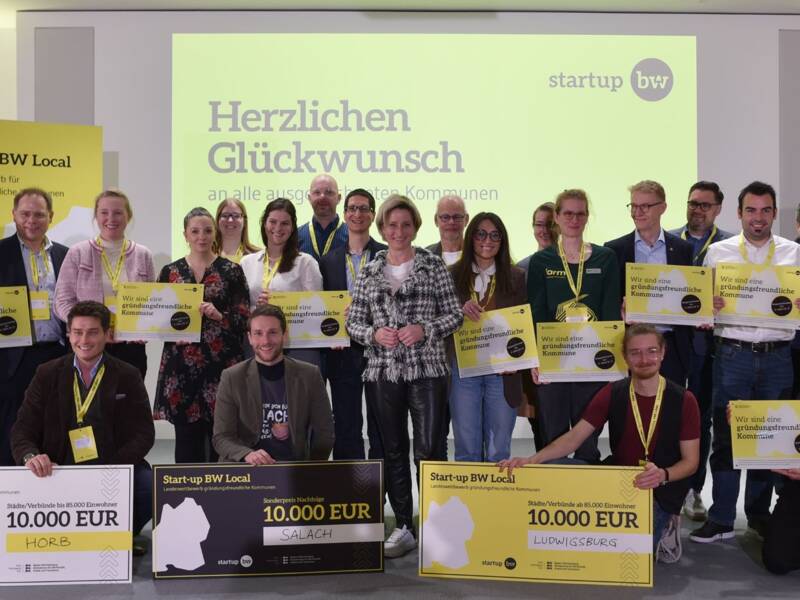 Eine Gruppe von Menschen hält auf einer Bühne verschiedene große Schecks und Schilder, während sie für ein Gruppenfoto vor einem gelb-weißen Hintergrund posiert, auf dem "Herzlichen Glückwunsch" steht.