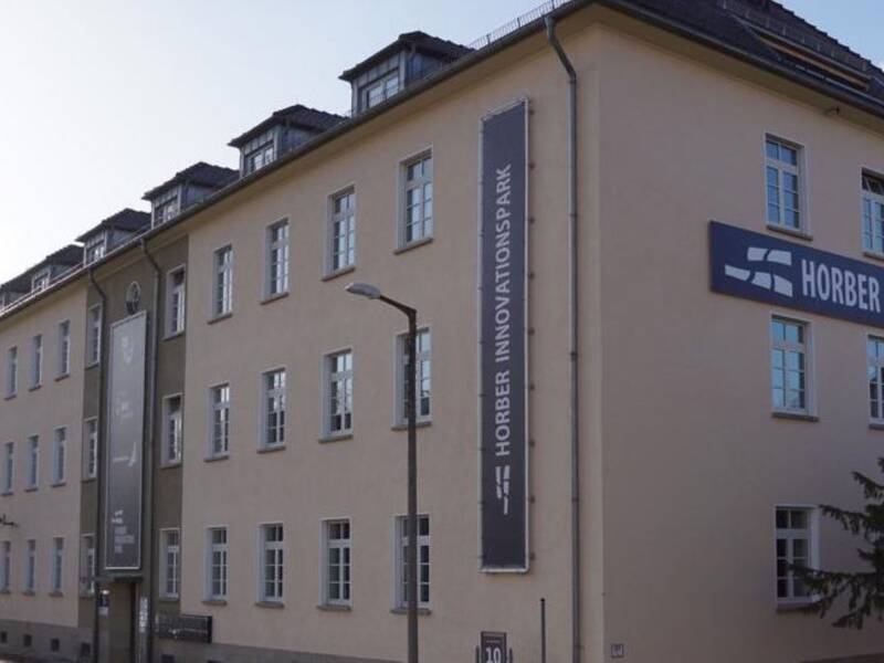Auf dem Bild ist ein Gebäude zu sehen. Auf der Kopfseite und auf der Straßenseite von dem Gebäude sind Banner mit dem Schriftzug Horber Innovationspark zu sehen. Auf der Kopfseite ist eine Tanne zu sehen und auf der Straßenseite vom Gebäude sind Straßenlaternen, ein Gehweg und eine Straße zu sehen. 