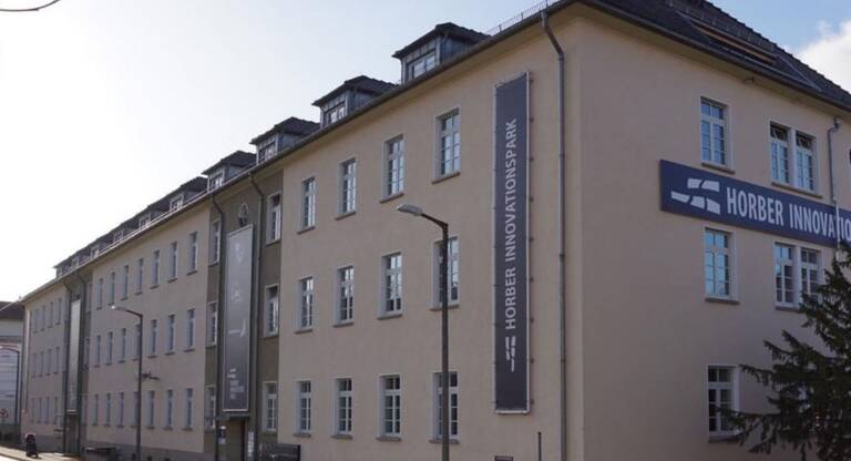 Auf dem Bild ist ein Gebäude zu sehen. Auf der Kopfseite und auf der Straßenseite von dem Gebäude sind Banner mit dem Schriftzug Horber Innovationspark zu sehen. Auf der Kopfseite ist eine Tanne zu sehen und auf der Straßenseite vom Gebäude sind Straßenlaternen, ein Gehweg und eine Straße zu sehen. 