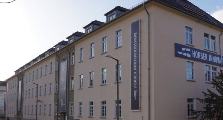 Auf dem Bild ist ein Gebäude zu sehen. Auf der Kopfseite und auf der Straßenseite von dem Gebäude sind Banner mit dem Schriftzug Horber Innovationspark zu sehen. Auf der Kopfseite ist eine Tanne zu sehen und auf der Straßenseite vom Gebäude sind Straßenlaternen, ein Gehweg und eine Straße zu sehen. 