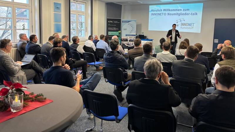 Ein Konferenzraum mit mehreren Personen, die in Richtung einer Präsentation sitzen, auf dem eine Wilkommensgruß zu sehen ist und der Moderator davor steht.