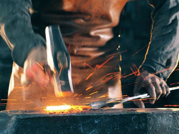 Ein Schmied hämmert mit einem großen Hammer auf ein glühendes Metallstück auf einem Amboss, wobei Funken sprühen.