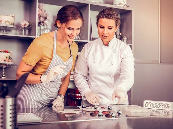 Zwei Frauen stehen in einer Küche und dekorieren Schokoladenpralinen, wobei die eine in einem weißen Kochanzug die andere, die ein gestreiftes Schürze trägt, anleitet.