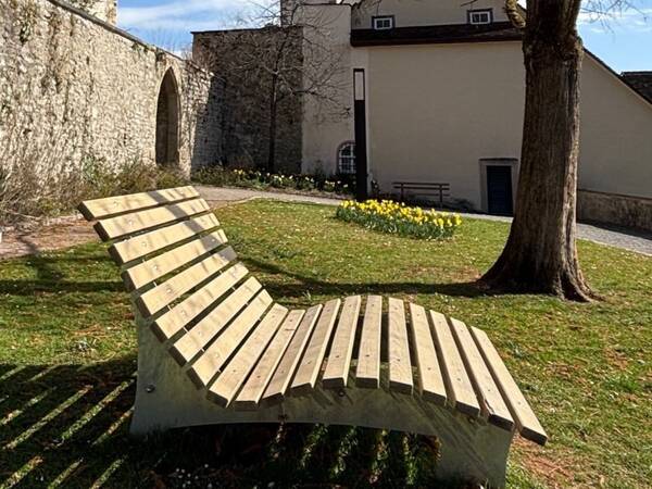 Eine Paarliege im Horber Burggarten vor einer alten Steinmauer und einem Gebäude im Hintergrund, während ein Baumstamm den Vordergrund schmückt.