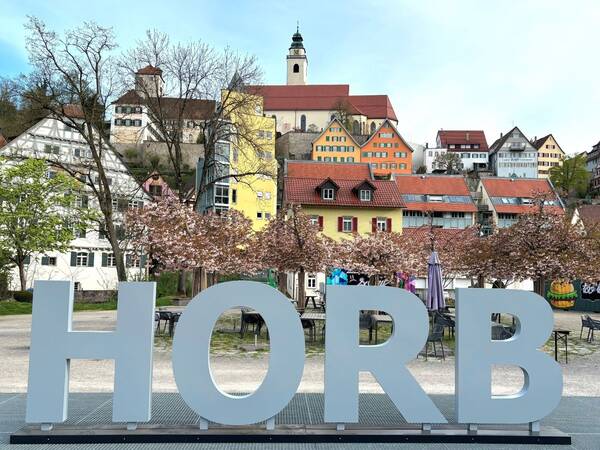 Eine große Skulptur mit dem Schriftzug "HORB" steht vor bunten Fachwerkhäusern und einer Kirche auf einem Hügel.