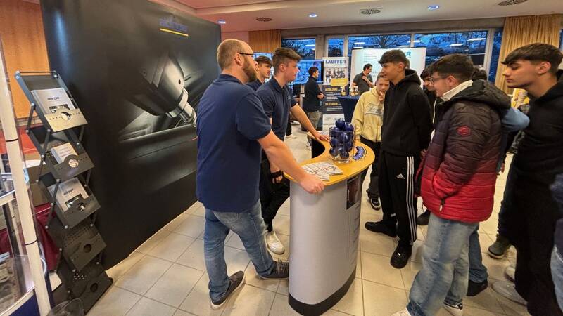 Das Bild zeigt Schülerinnen und Schüler, die im Gespräch sind und an einem Messestanden stehen. Im Hintergrund ist ein weiterer Messestand zu sehen. 