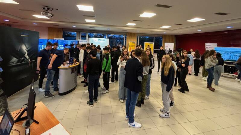 Das Bild zeigt ein Foyer mit verschiedenen Messeständen im Hintergrund. Im Vordergrund sind Schülerinnen und Schüler, die im Gespräch sind und an Messeständen stehen. 