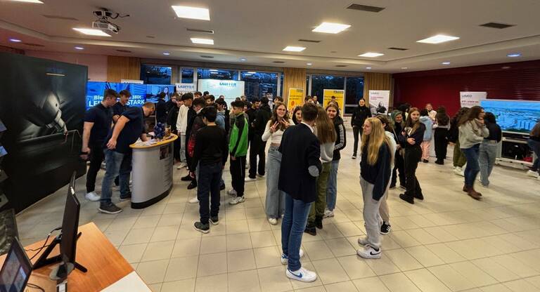 Das Bild zeigt ein Foyer mit verschiedenen Messeständen im Hintergrund. Im Vordergrund sind Schülerinnen und Schüler, die im Gespräch sind und an Messeständen stehen. 