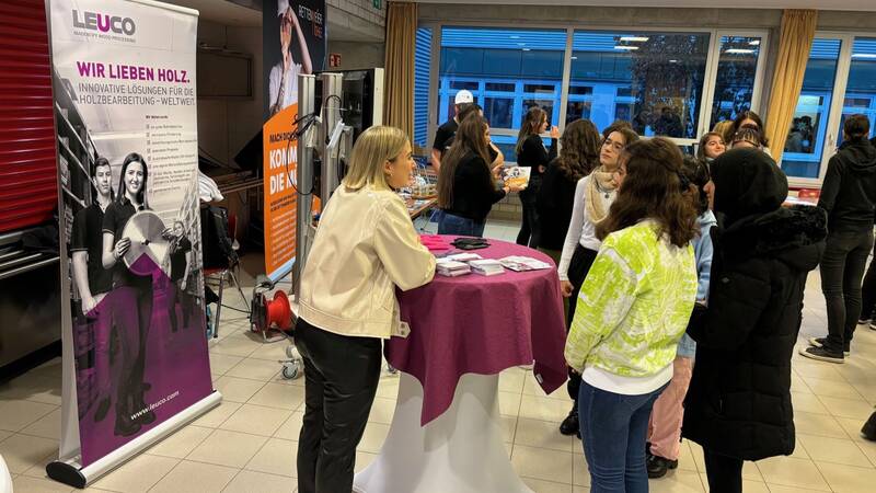 Das Bild zeigt Schülerinnen, die im Gespräch sind und an einem Messestanden stehen. Im Hintergrund ist ein weiterer Messestand, Schülerinnen und Schüler zu sehen. 