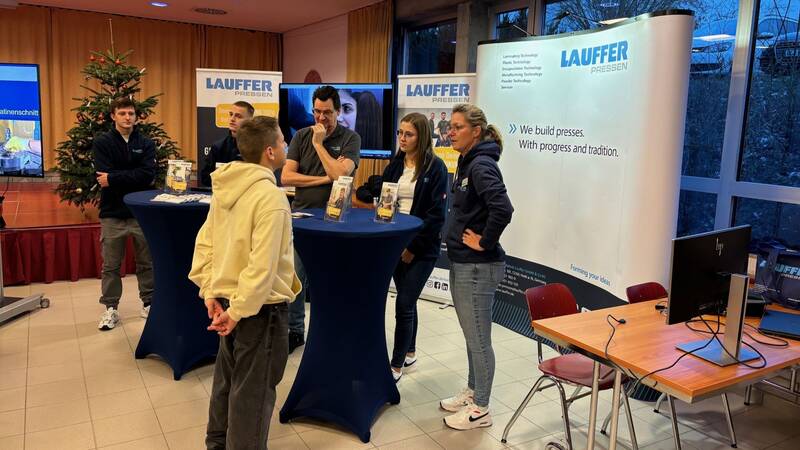 Das Bild zeigt einen Schüler, der an einem Messestand steht und sich mit den Messestandpersonal im Gespräch befindet. Im Hintergrund sind zwei weitere Personen von dem Messepersonal, eine Bühne auf der ein Weihnachtsbaum steht und ein Stück von einem Fernseher. 