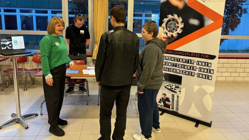 Das Bild zeigt zwei Schüler, die an einem Messestanden stehen und sich im Gespräch mit einer Frau von dem Messestand befinden. Im Hintergrund ist eine weitere Person von dem Messestand, Stühle und Fenster zu sehen.