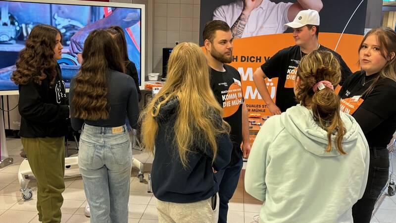 Das Bild zeigt Schülerinnen, die an einem Messestanden stehen und sich im Gespräch mit dem Messestandpersonal befinden. Im Hintergrund ist eine Roll-Up Banner und ein Fernseher zu sehen. 