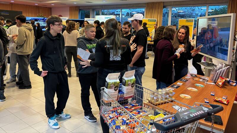 Das Bild zeigt ein Foyer mit verschiedenen Messeständen im Hintergrund. Im Vordergrund sind Schülerinnen und Schüler, die im Gespräch sind und an Messeständen stehen. 