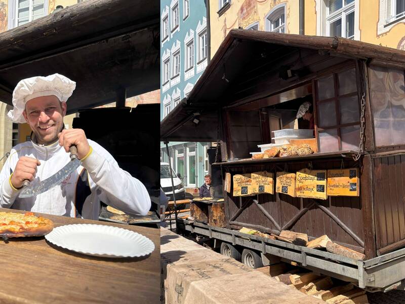 Auf dem Bild sieht man rechts einen Holzwagen, in dem Essen verkauft wird. Im Hintergrund sieht man bunte Gebäude und Menschen. Auf der linken Seite ist ein lächelnder Mann zu sehen mit einem Schneidemesser in der Hand.