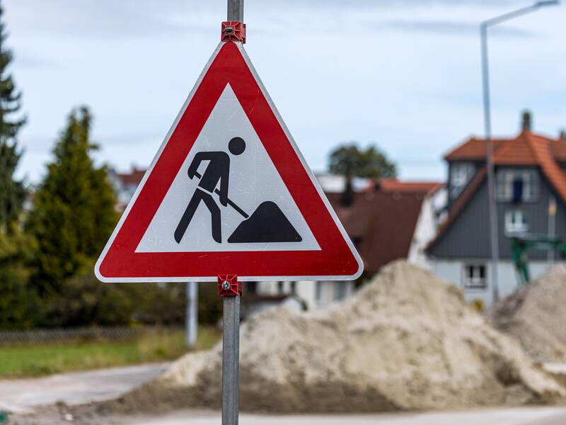 Ein Verkehrsschild, das auf eine Baustelle hinweist. Im Hintergrund befinden sich Sandhäufen auf einer Straße.