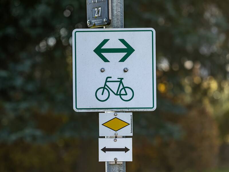 Ein weißes Schild mit einem grünen Fahrrad und Richtungspfeil in beider Richtungen als Wegweiser für einen Radweg. Das Schild ist an einem Laternenpfosten befestigt.